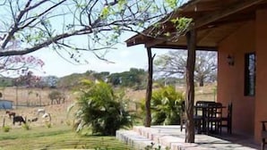 Un bâtiment doté d’un patio couvert, d’un espace extérieur aménagé pour s’asseoir et offrant une vue sur un champ verdoyant, avec des arbres et une clôture en arrière-plan.