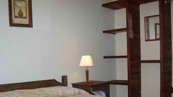 A bedroom with a bed, a bedside table with a lamp, a framed picture on the wall, and a wooden shelf unit.