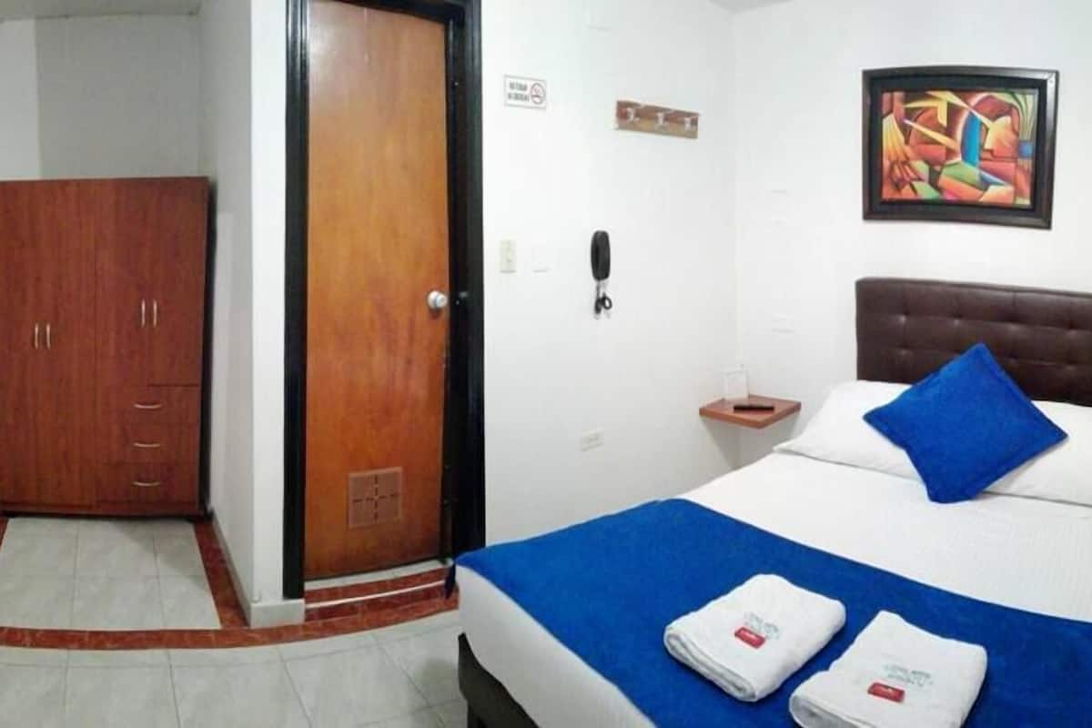 Habitación de hotel con cama, armario de madera, puerta, un cuadro en la pared y un teléfono en la mesita de noche.
