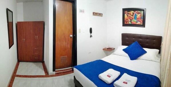 Quarto de hotel com cama, guarda-roupa de madeira, porta, quadro na parede e telefone na mesinha de cabeceira.