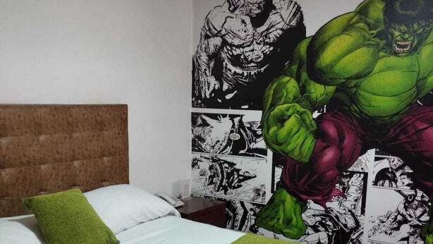 A hotel room with a large bed, green pillows, a green throw blanket, and a wall mural featuring a green muscular character. - Hotel Comic City (Bogotá)