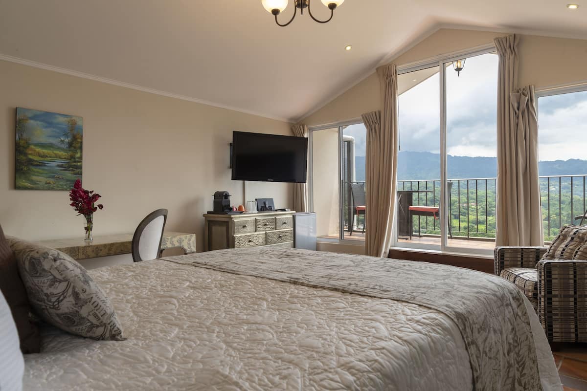 Un dormitorio con una cama grande, un televisor montado en la pared, un escritorio con una computadora y vistas a montañas a través de amplios ventanales.