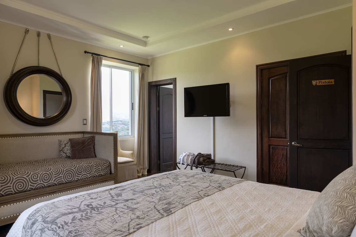 Habitación de hotel con una cama grande, un sofá más pequeño, un espejo redondo, un televisor y una puerta con un letrero.