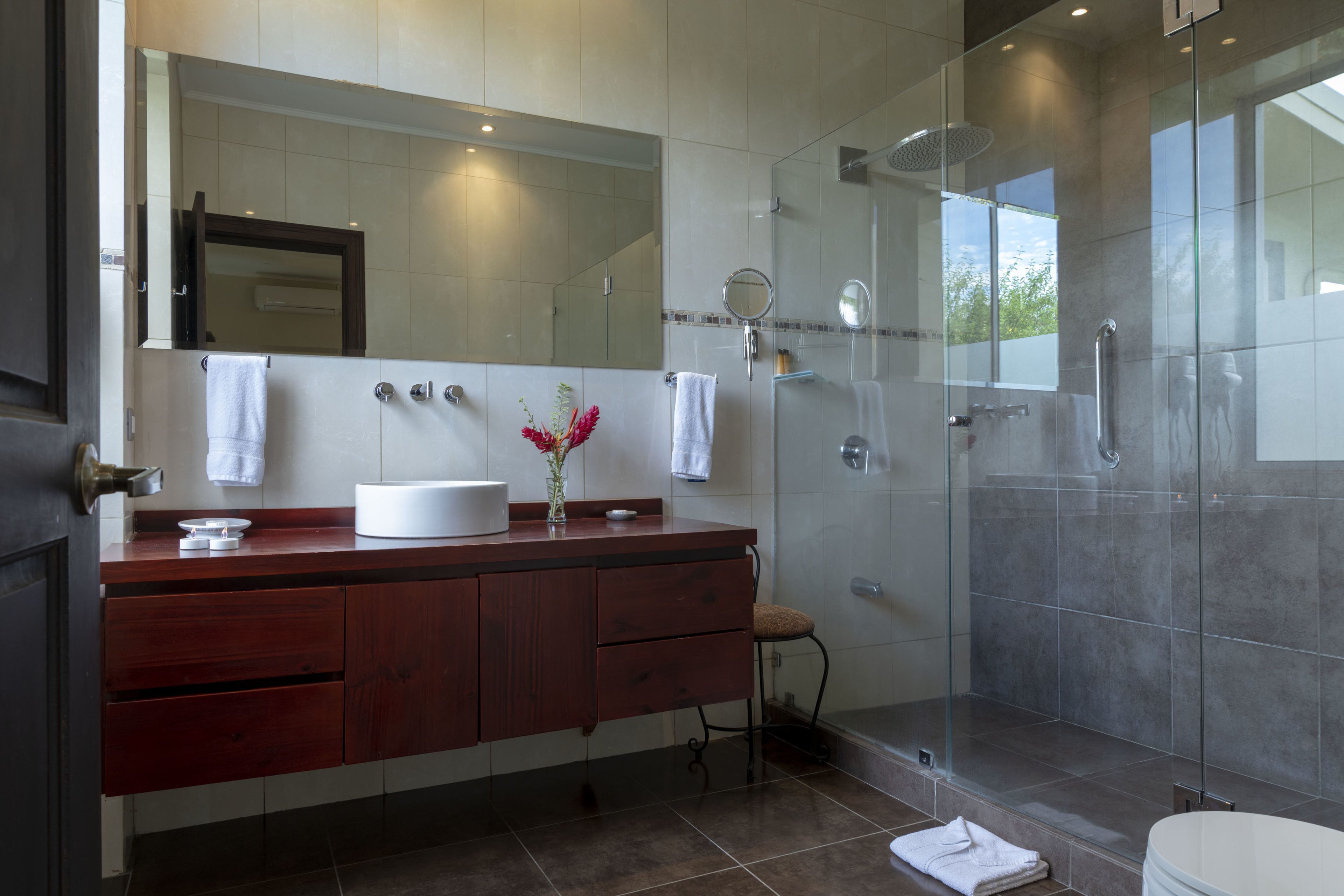 Une salle de bain moderne dotée d’une douche à parois de verre, d’un grand miroir et d’un meuble-lavabo en bois avec un évier et un porte-serviettes.