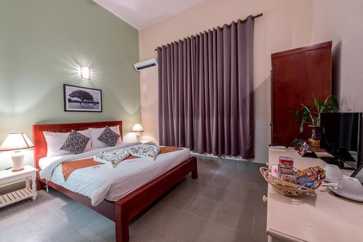 Cama bem arrumada com almofadas, abajur, quadro na parede, janela com cortinas e uma pequena mesa com objetos.