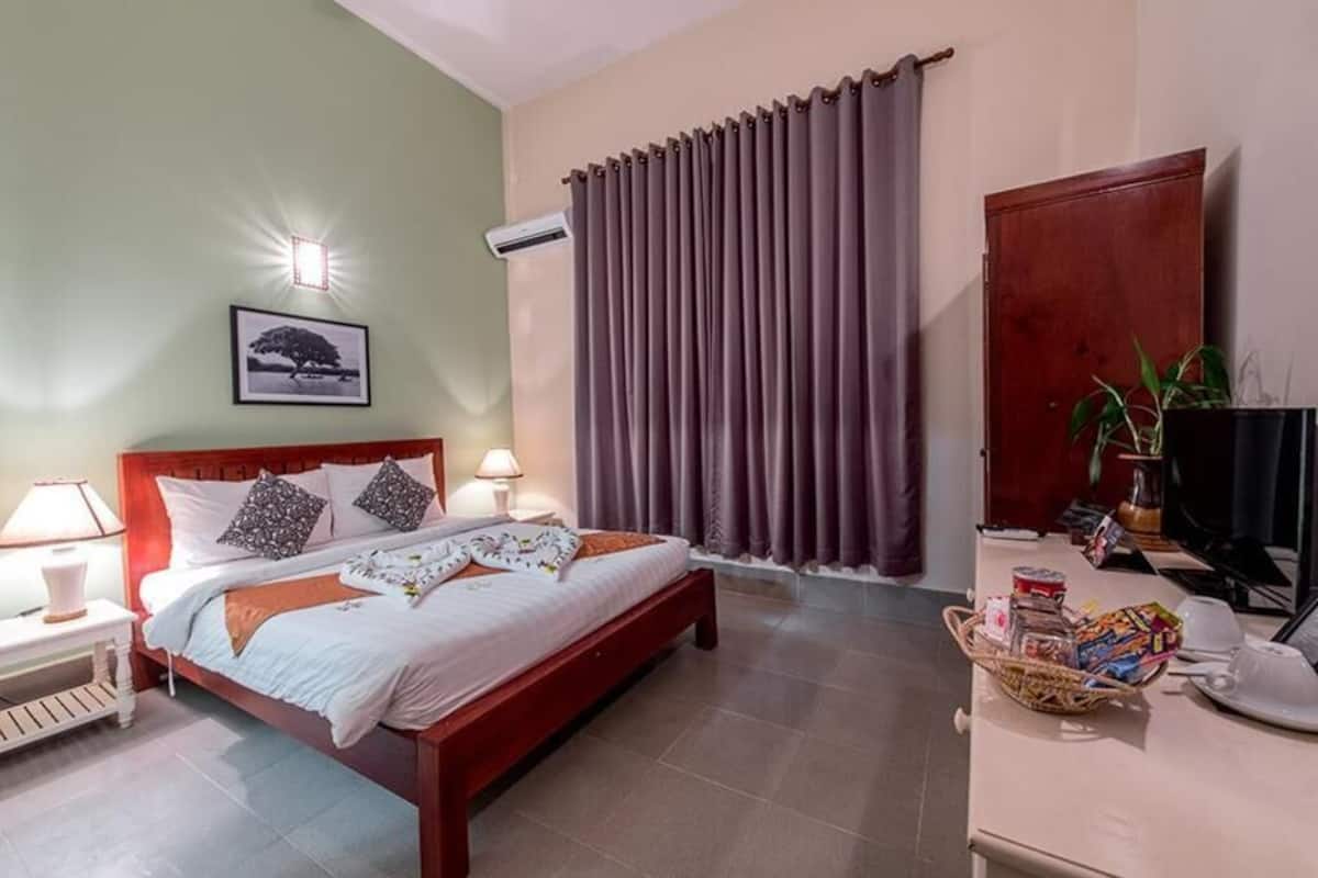 A neatly made bed with pillows, a bedside lamp, a framed picture on the wall, a window with curtains, and a small table with items.