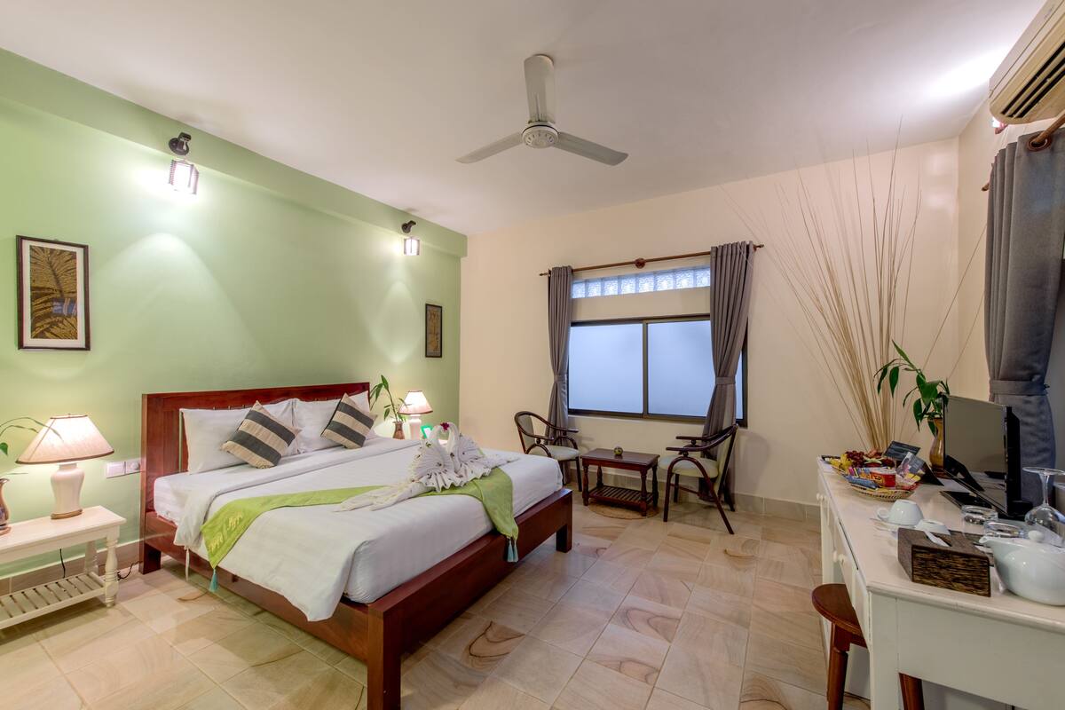Quarto de hotel com cama, mesas de cabeceira, escrivaninha e cadeira, ventoinha no teto e janela com cortinas.