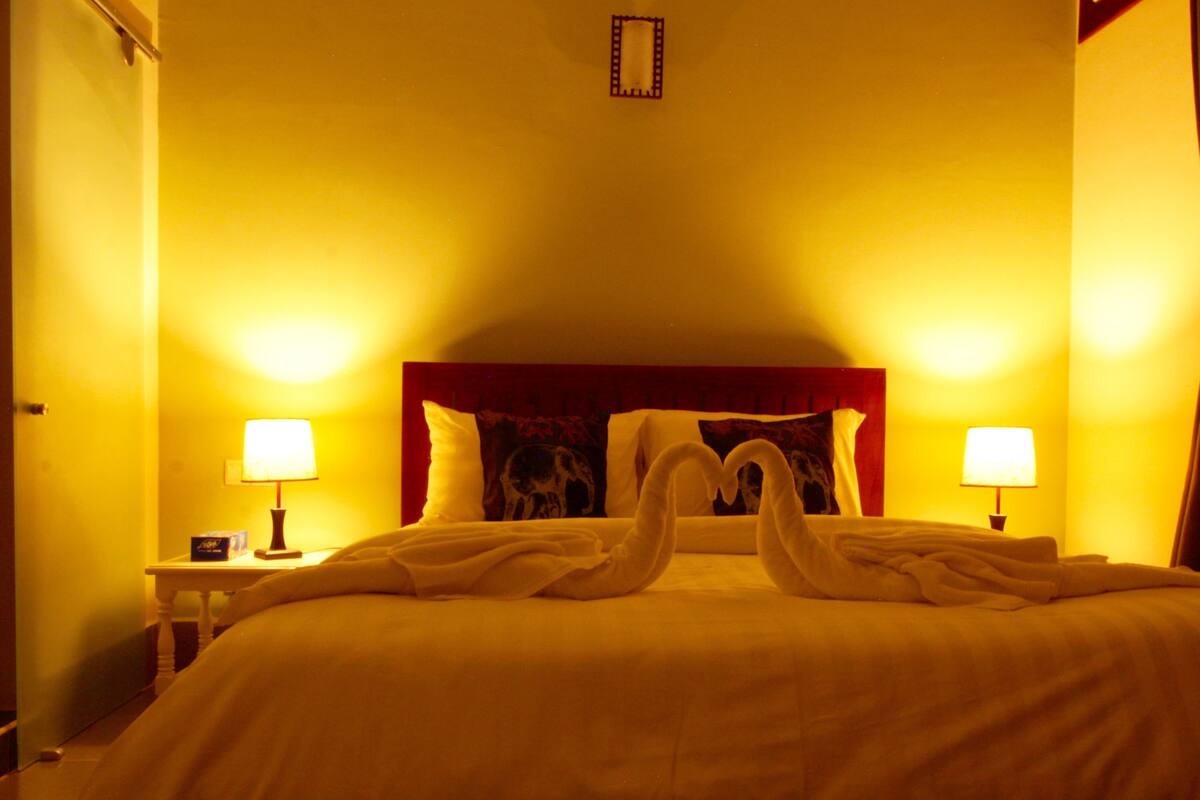 Quarto de hotel com cama, duas luminárias de cabeceira, painel de cabeceira e luminária fixa na parede.