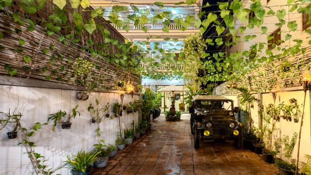 Une ruelle étroite où est garée une vieille jeep sur le côté droit, entourée d’une végétation luxuriante et de plantes en pot. On peut apercevoir, au fond, une enseigne indiquant l’hôtel « Lemongrass & Ginger ».