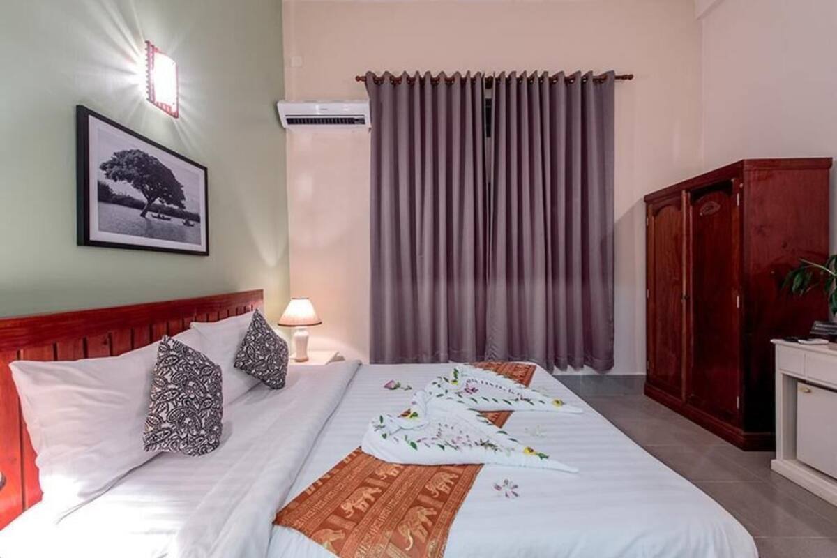 Uma cama bem arrumada com almofadas, um abajur, um quadro na parede e uma janela com cortinas.