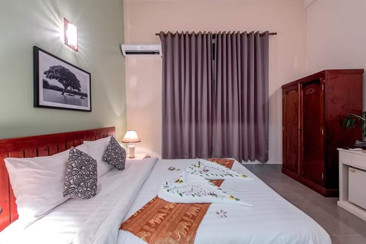 A neatly made bed with pillows, a bedside lamp, a framed picture on the wall, and a window with curtains.