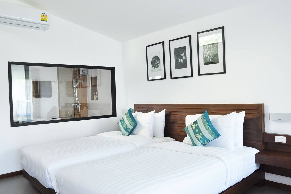 Une chambre d’hôtel avec un grand lit, une tête de lit en bois et une grande fenêtre. Trois tableaux encadrés sont accrochés au mur, au-dessus du lit.