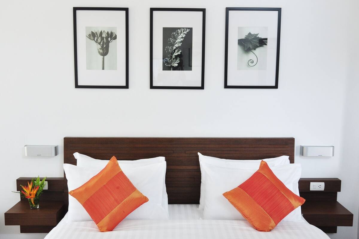 Un lit bien fait, agrémenté d’oreillers blancs et orange, d’une tête de lit en bois foncé et de trois reproductions botaniques encadrées accrochées au mur au-dessus.