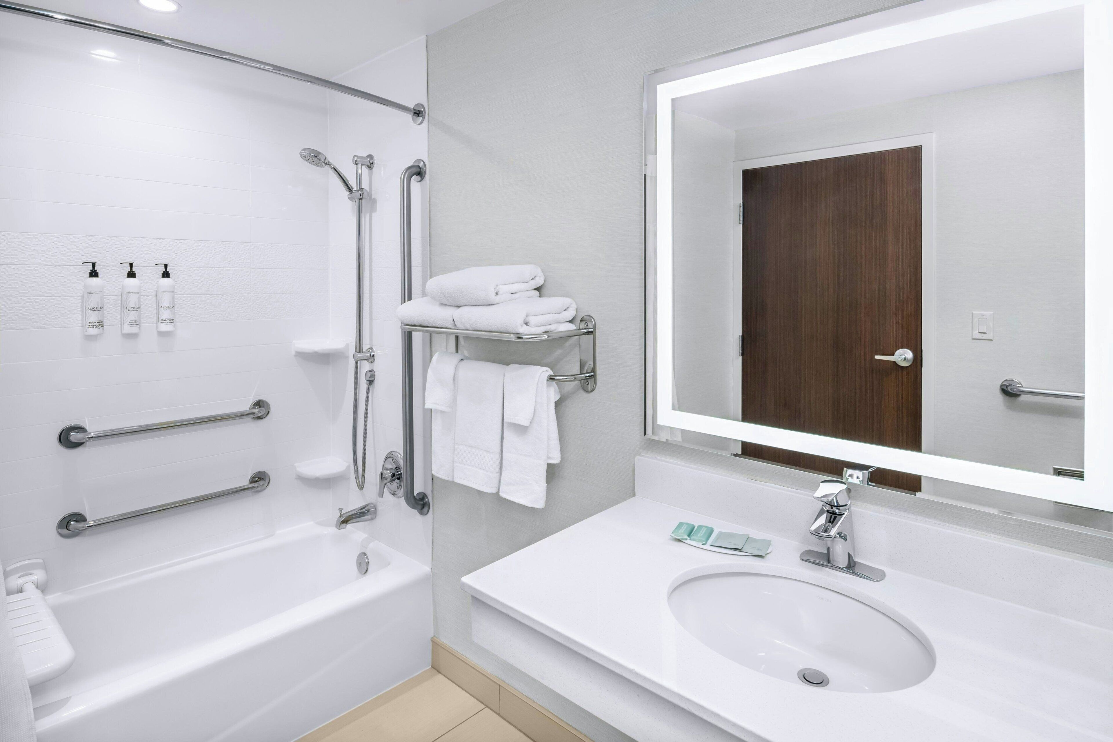 Een moderne badkamer met een badkuip, douche en wastafel. Er zijn handgrepen, een handdoekrek en een spiegel boven de wastafel.