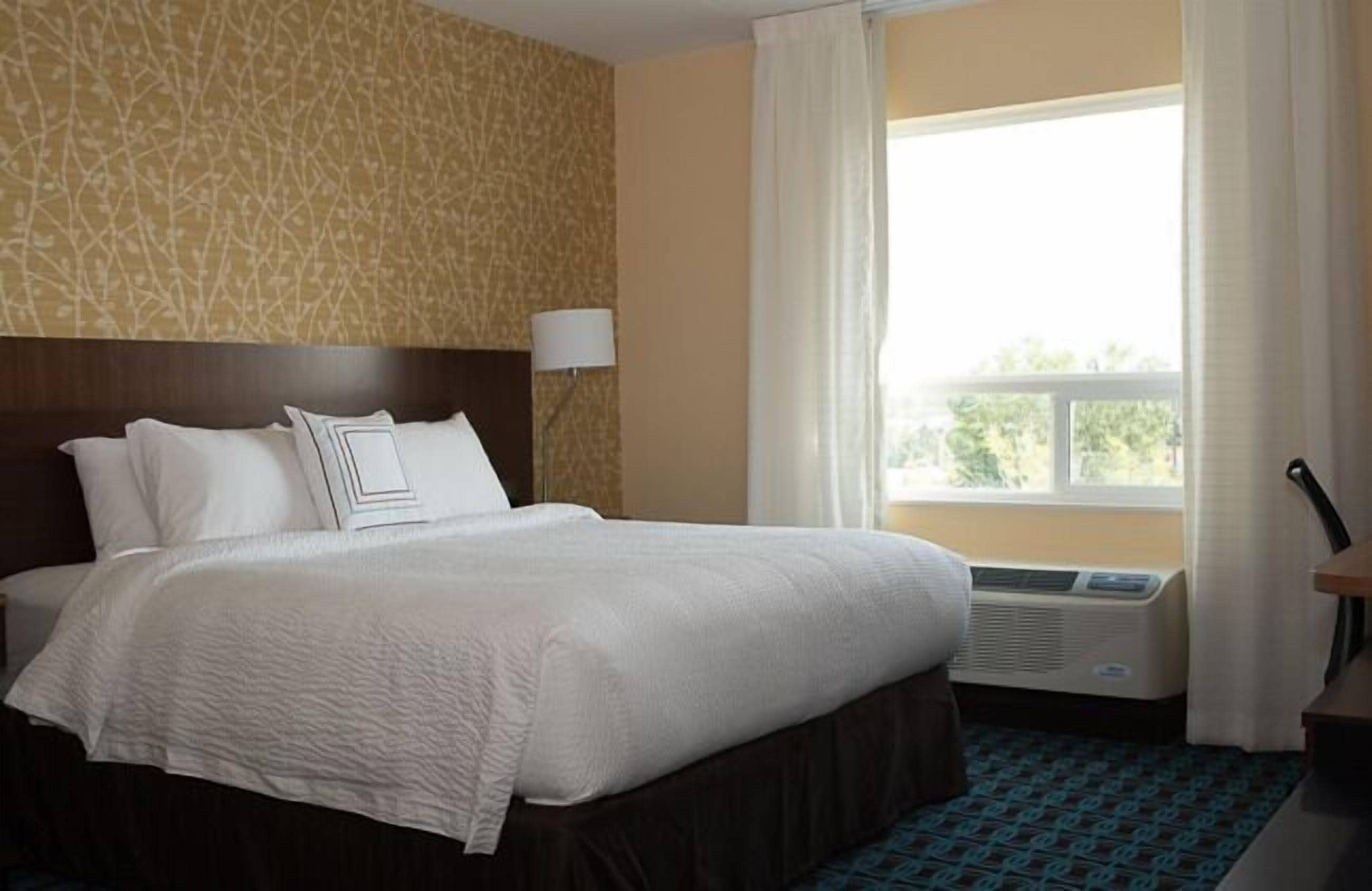 Une chambre d’hôtel avec un grand lit, une tête de lit, une fenêtre avec des rideaux, un appareil de climatisation et une moquette à motifs.