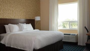 A hotel room with a large bed, a headboard, a window with curtains, an air conditioning unit, and a patterned carpet.