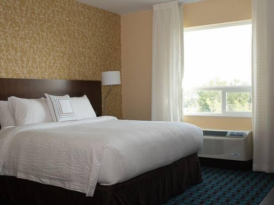 A hotel room with a large bed, a headboard, a window with curtains, an air conditioning unit, and a patterned carpet.