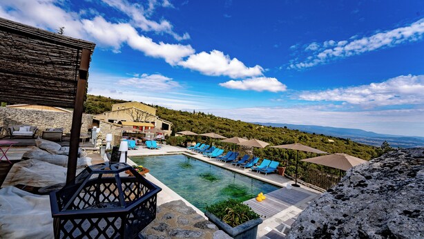 A pool area with lounge chairs and umbrellas, a stone building, and a view of the landscape.