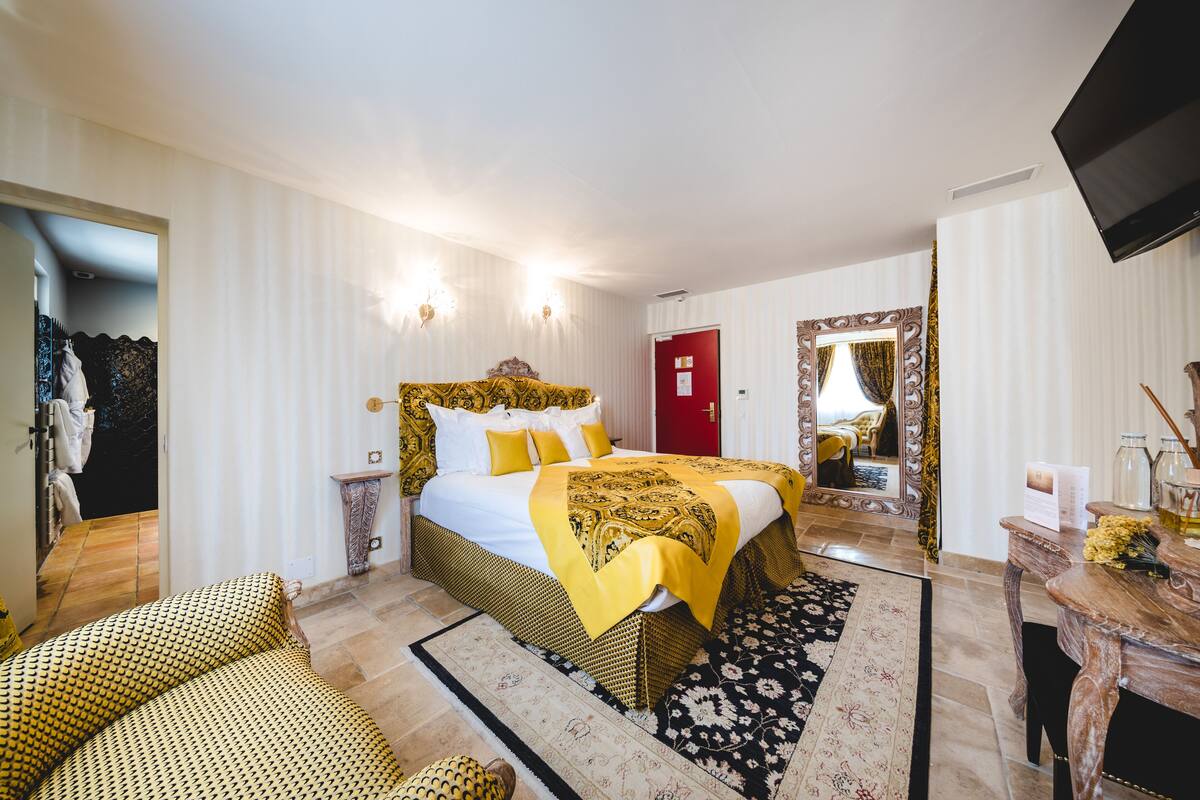Une chambre d’hôtel avec un grand lit, une tête de lit en bois, une porte rouge, un téléviseur et une petite table sur laquelle est posé un vase de fleurs.