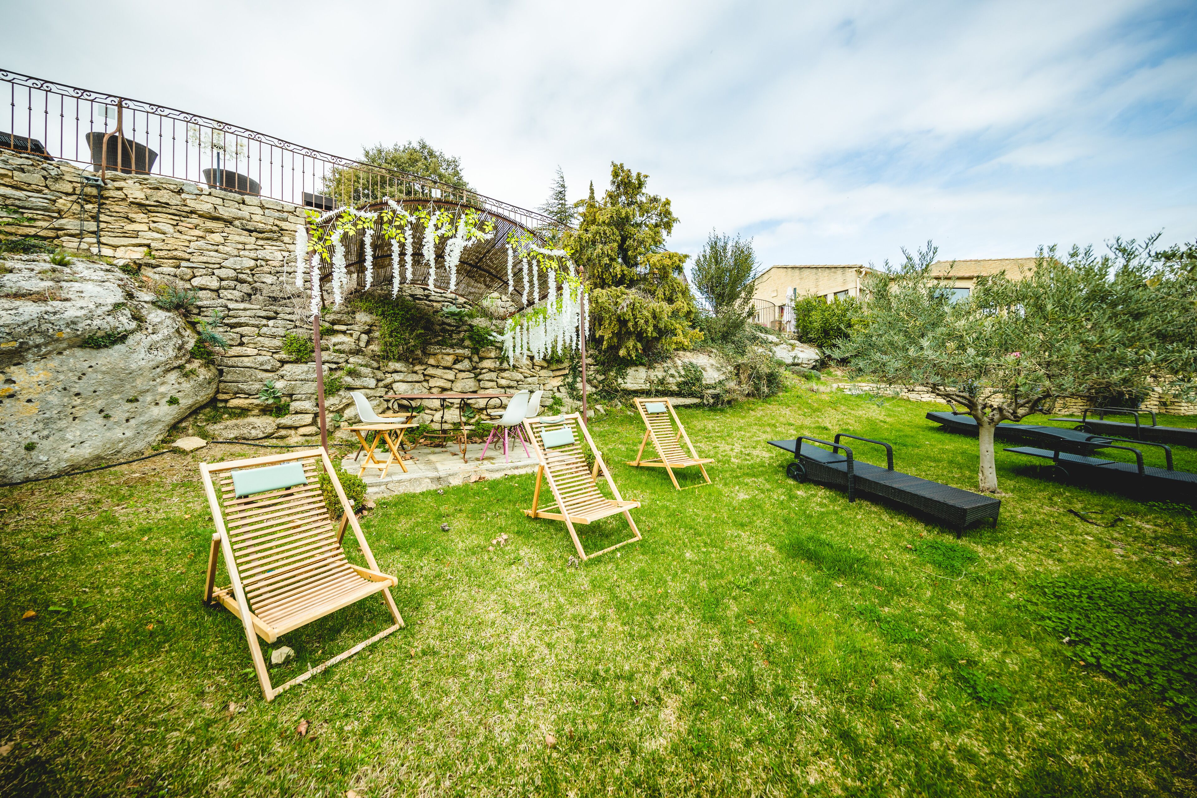 A stone wall, wooden lounge chairs, and a grassy area with a metal fence.