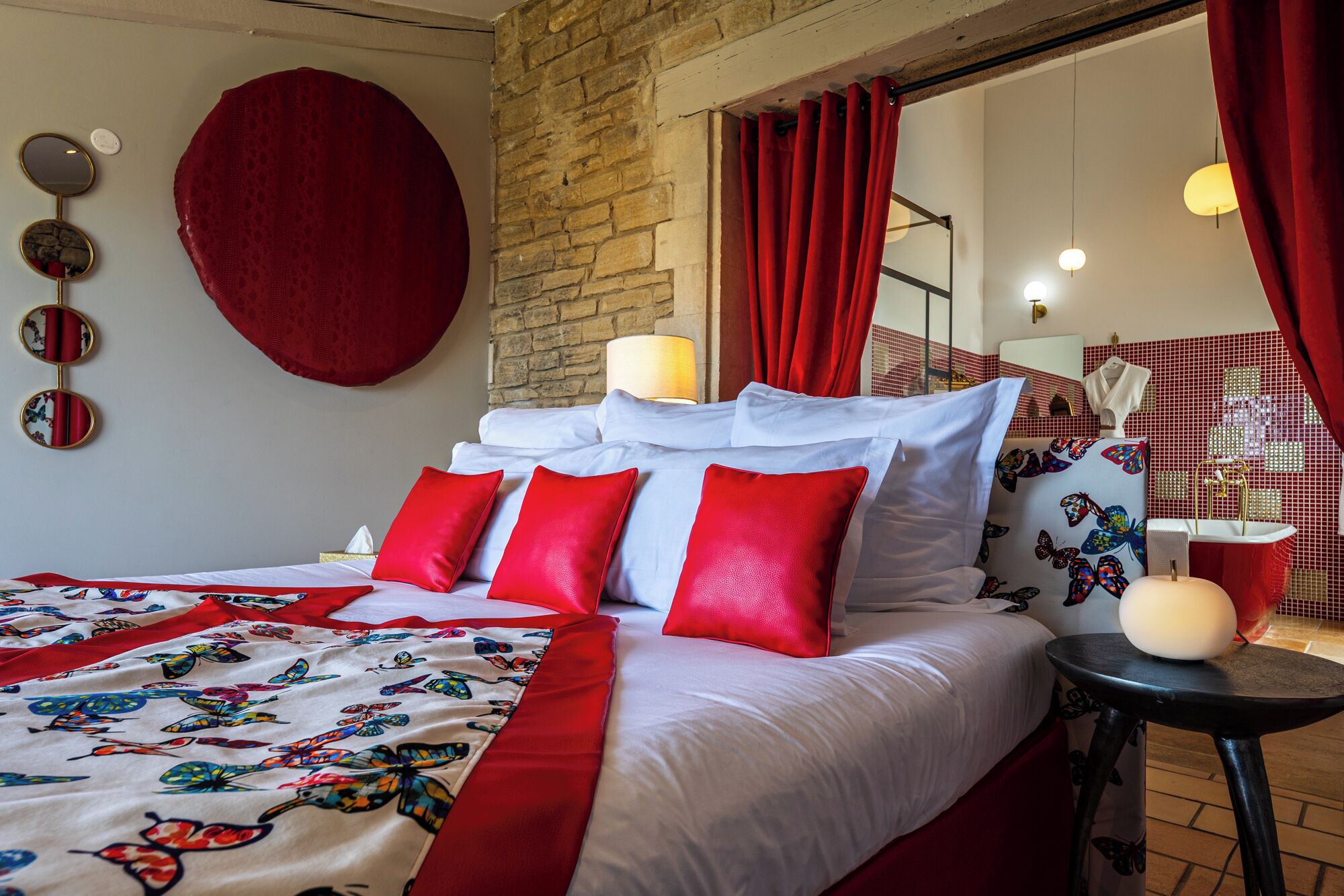 Een hotelkamer met een groot bed, rode kussens, een bedspreide met vlinderafdruk, een ronde spiegel, een rood gordijn en een badkamer met een douchegordijn met vlinders.