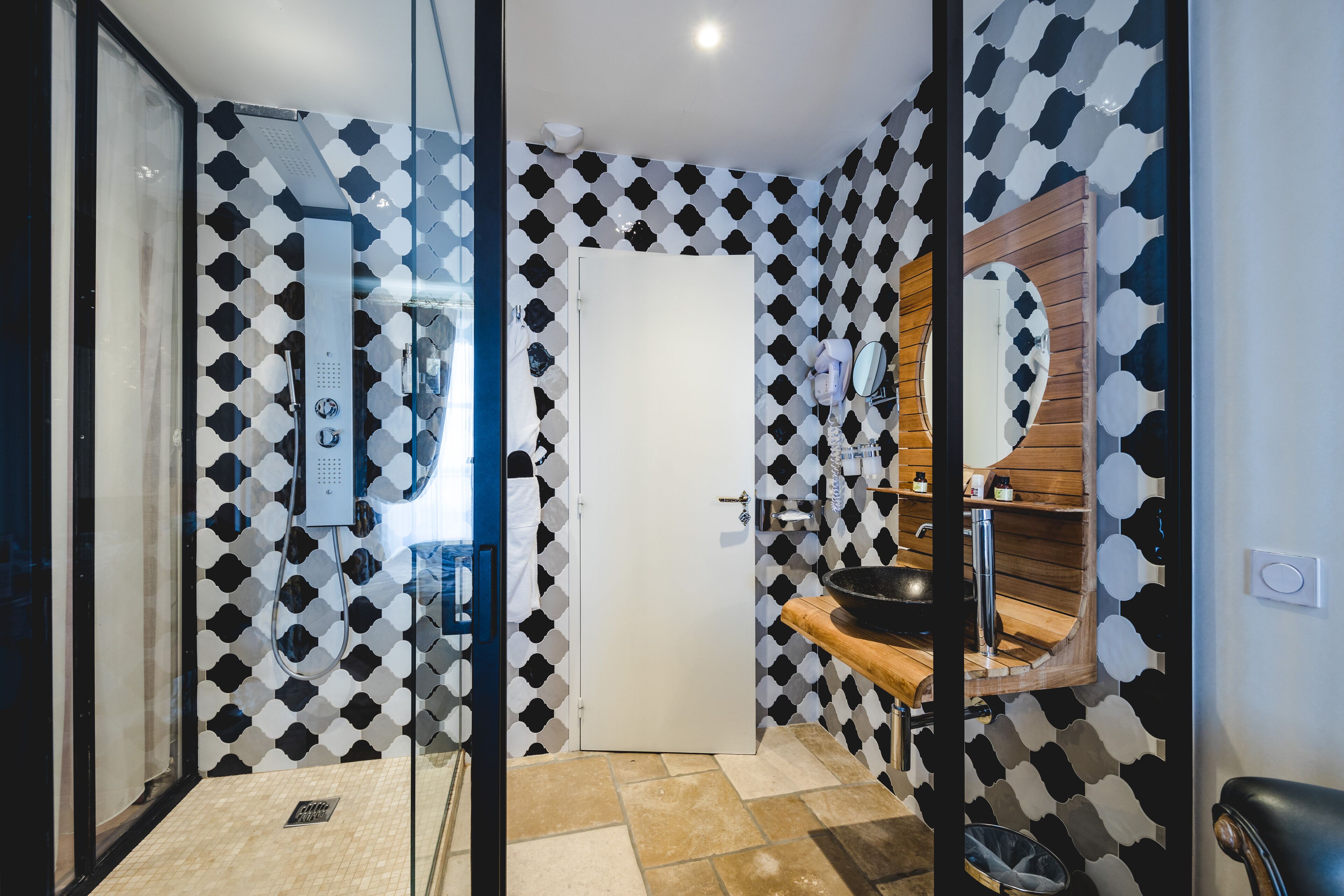 A modern bathroom with a geometric patterned wall, a glass shower enclosure, a wooden vanity, and a large mirror.