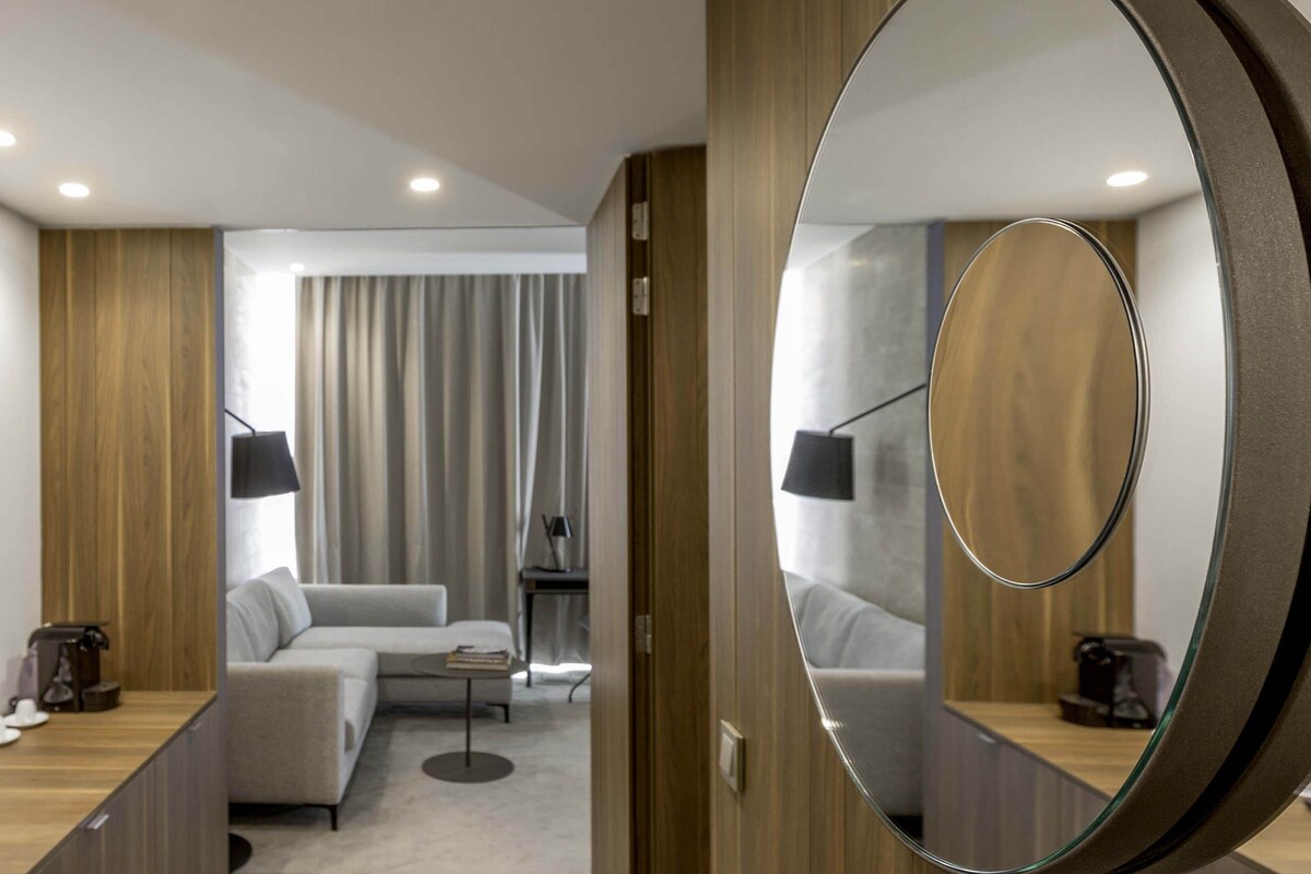 A modern hotel room with a sofa, a coffee table, a mirror, and a desk with a lamp.