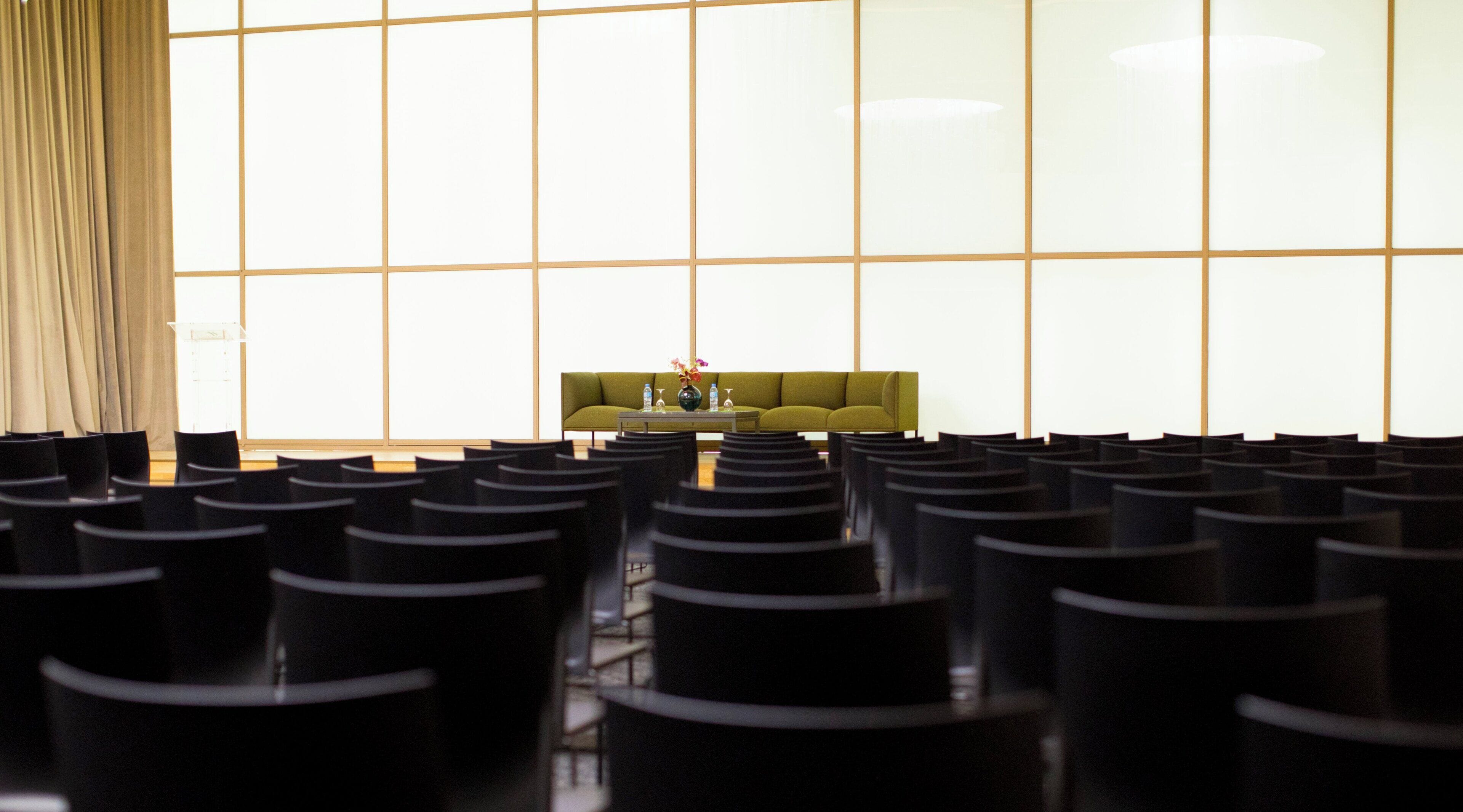 Un salón de conferencias con filas de sillas orientadas a un atril, un sofá verde y un amplio ventanal con diseño de cuadrícula.