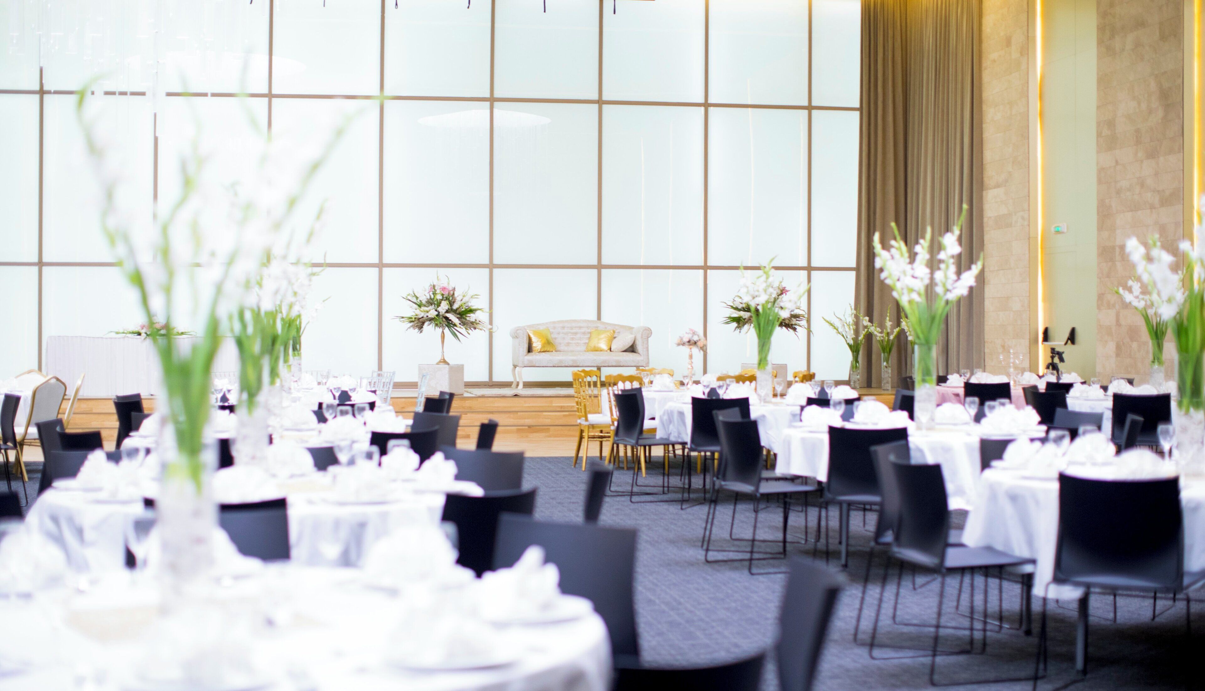 A banquet hall with round tables set for dining, a stage with a sofa, and floral centerpieces.