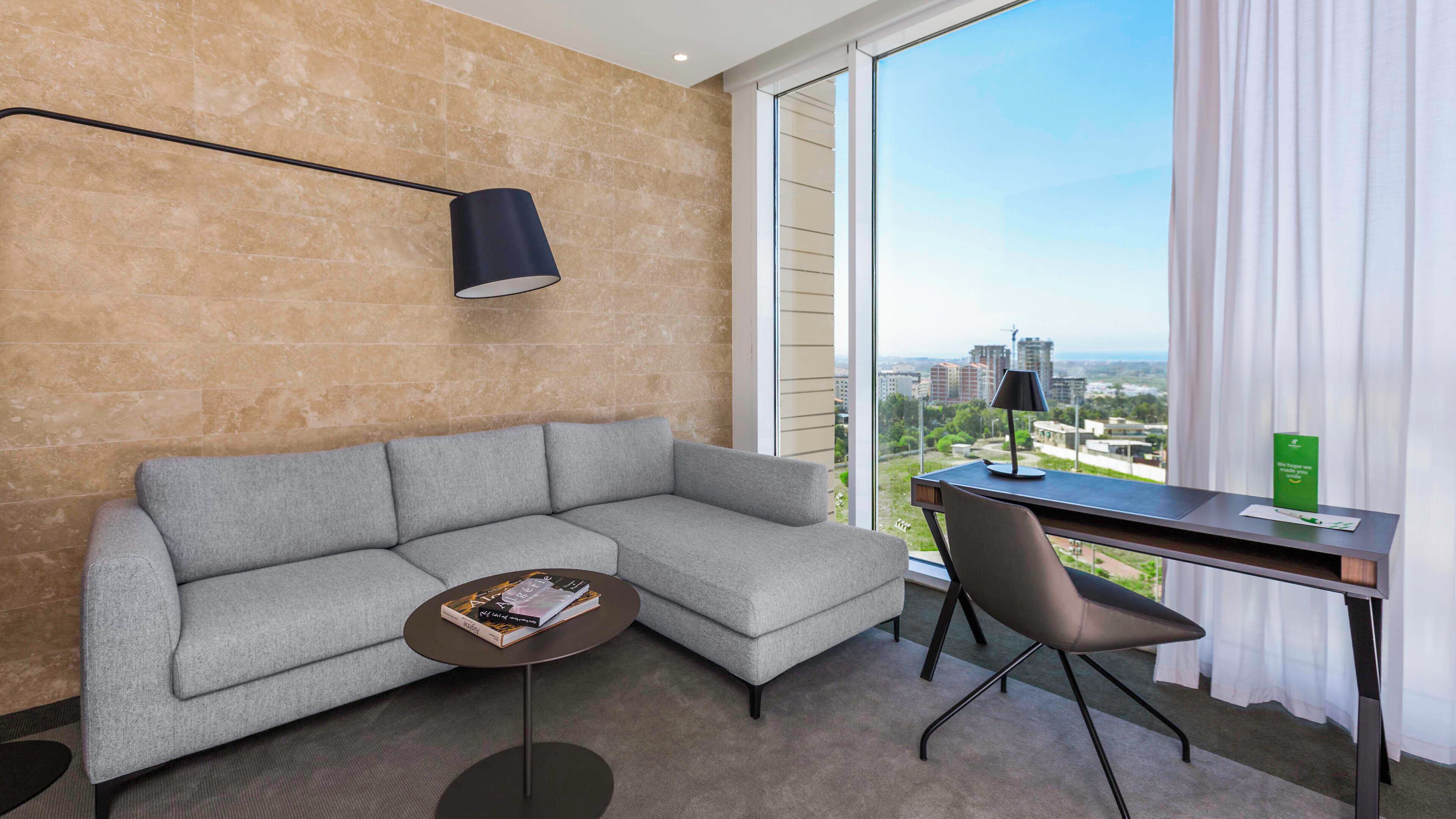 Habitación de hotel moderna con un sofá gris, una mesita redonda con un libro, un escritorio con una silla y vistas a la ciudad a través de grandes ventanales.