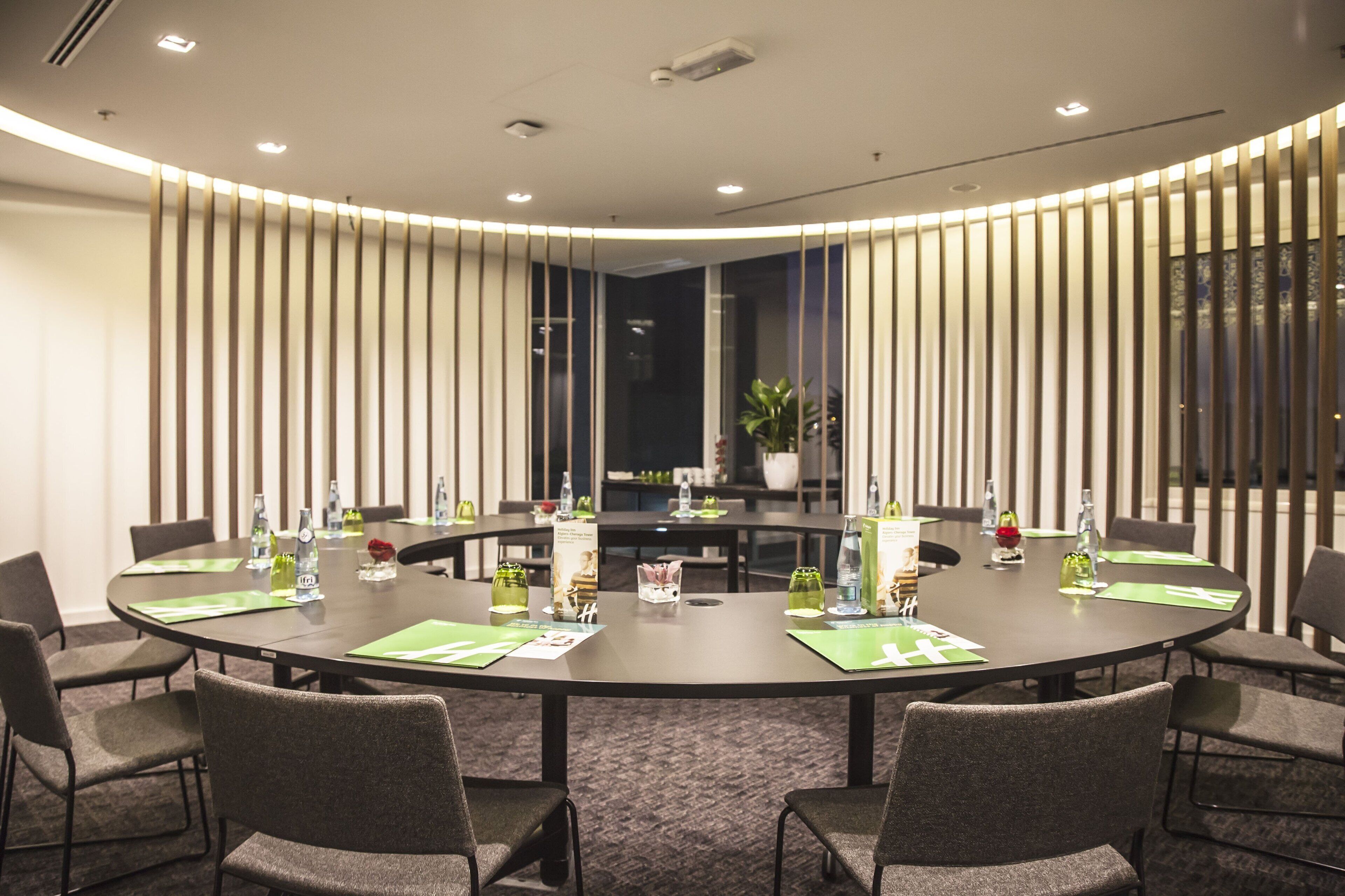 Un salón de conferencias con una mesa redonda, sillas y varios elementos como botellas de agua y un jarrón con flores.