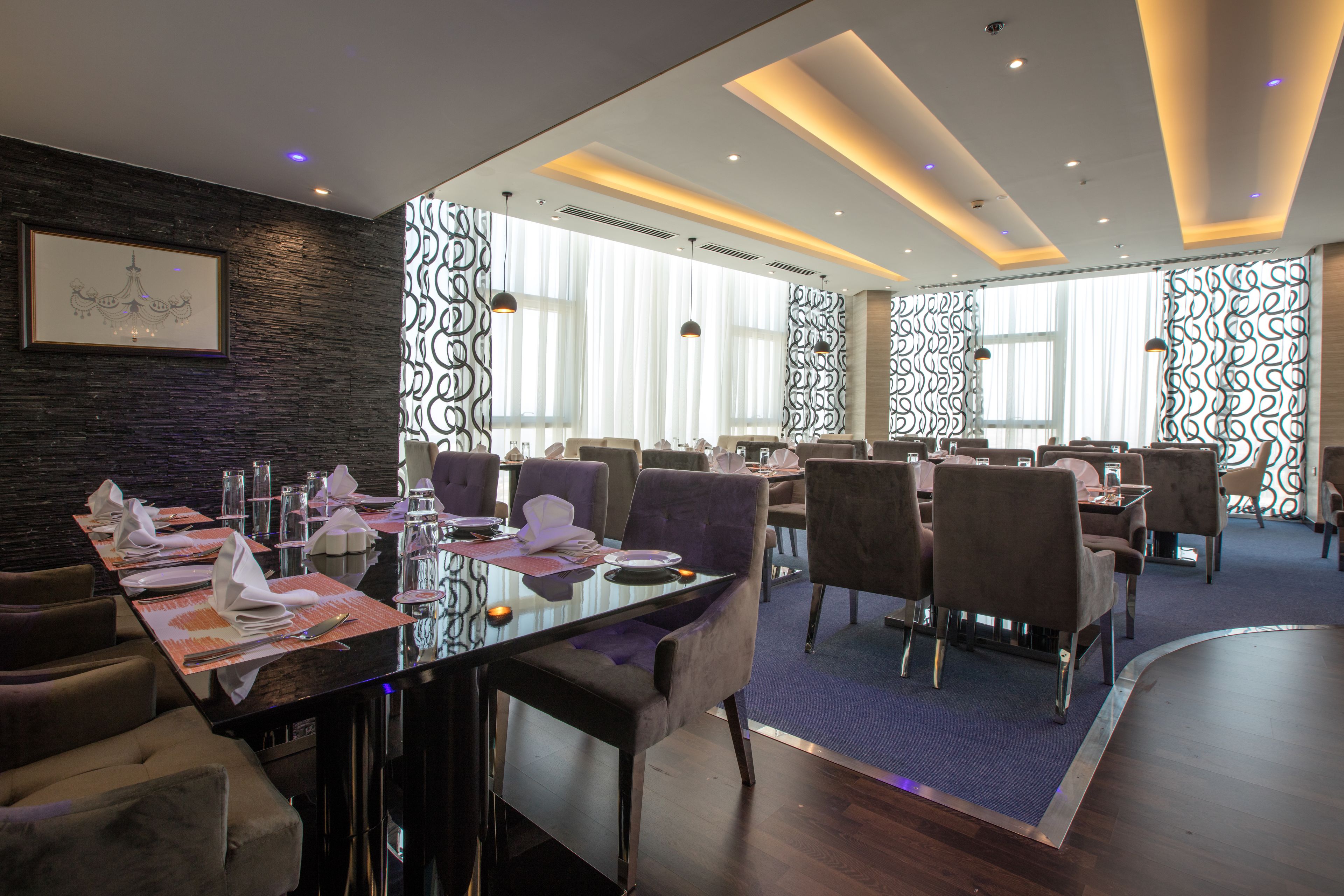 A modern dining area with a long table set for a meal, surrounded by comfortable chairs, and large windows providing natural light.