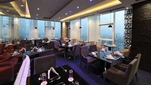 Un restaurante moderno con mesas y sillas de vidrio, ventanas amplias con cortinas y vistas a la ciudad.