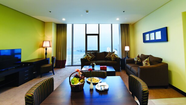 Una habitación de hotel moderna con un ventanal grande, un televisor de pantalla plana, un sofá, una mesa de centro y una mesa de comedor preparada con fruta.