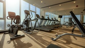 Un gimnasio con cintas de correr, elípticas y pesas libres, amplias ventanas y vista a la ciudad.