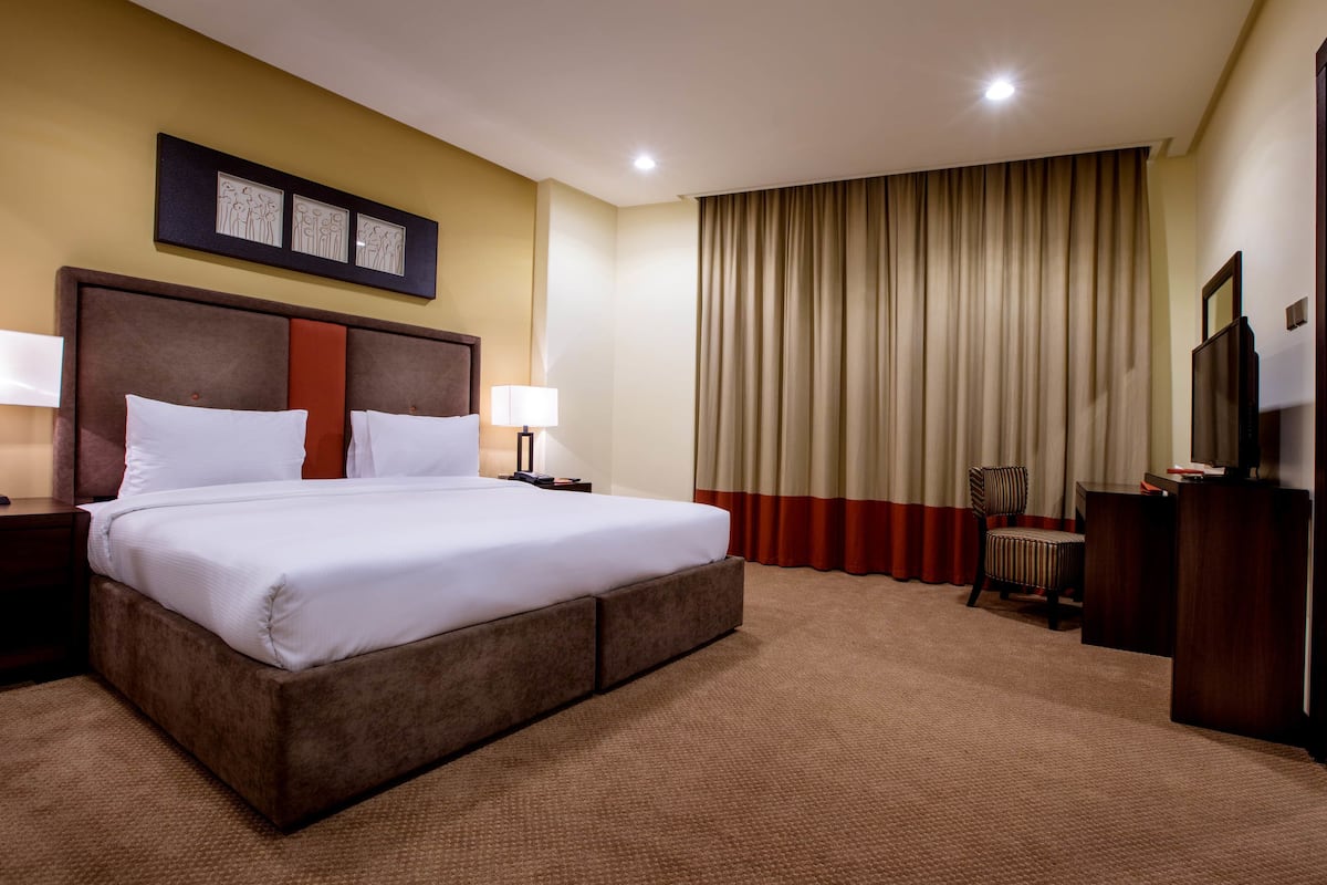 Une chambre d’hôtel avec un grand lit, deux tables de chevet avec des lampes, un bureau, une chaise et une grande fenêtre avec des rideaux.