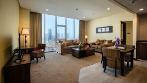 Una habitación de hotel moderna con un ventanal amplio que ofrece vistas a la ciudad, una zona de estar acogedora con un sofá y sillones, una pequeña mesa de comedor y un escritorio con lámpara.