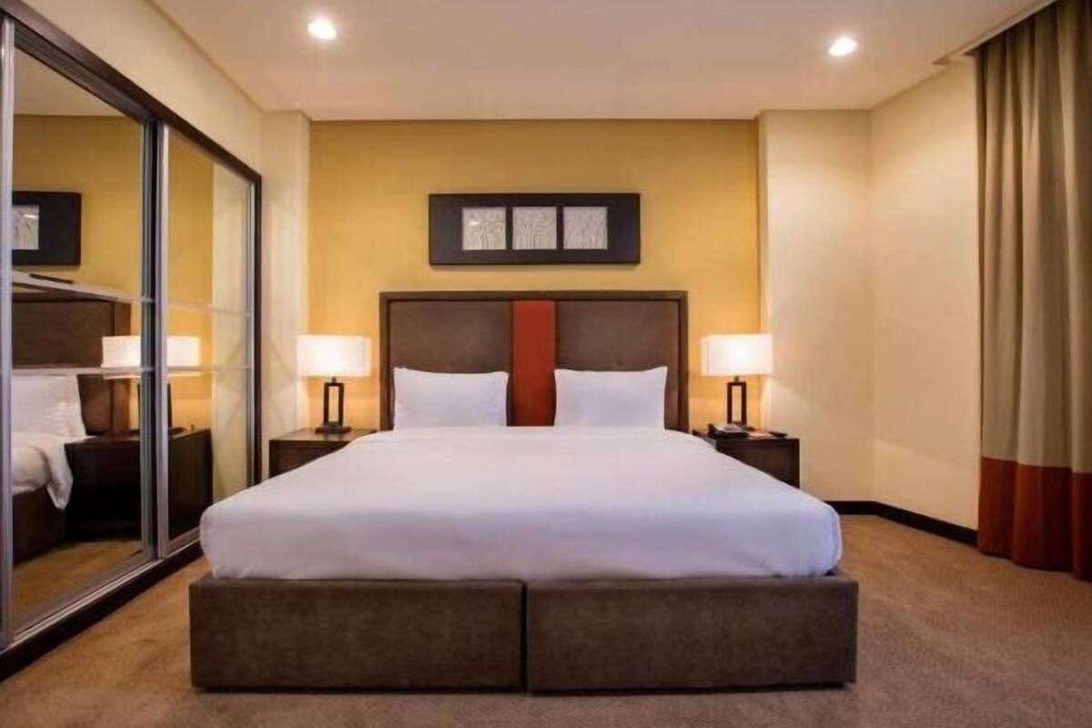 Une chambre d’hôtel avec un grand lit, deux tables de chevet avec des lampes, une tête de lit agrémentée d’une touche de rouge et un miroir sur un des murs.