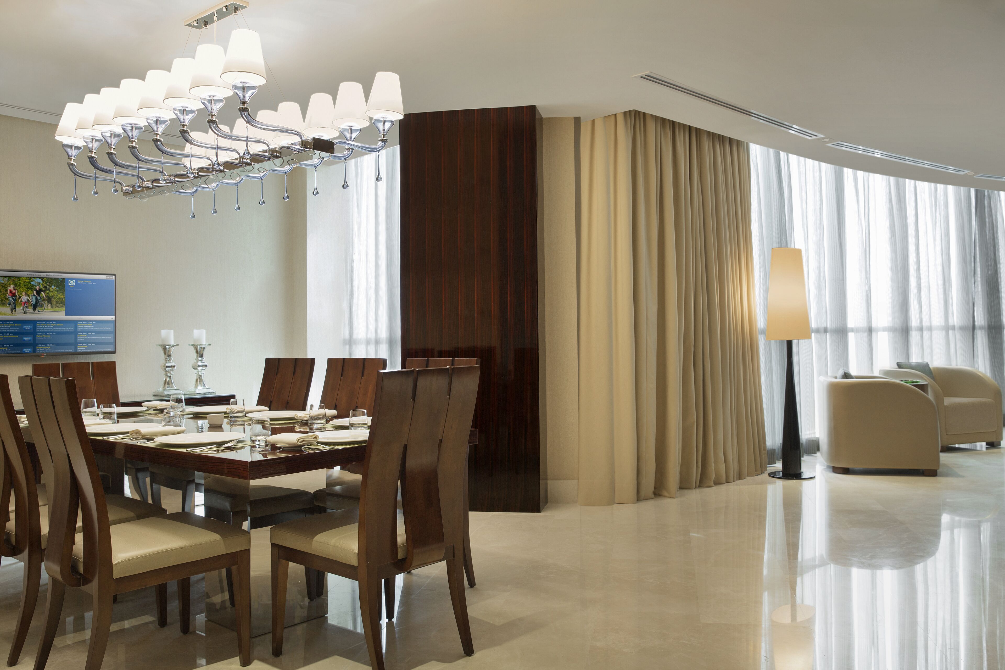 A modern hotel dining area with a large wooden table set for six, surrounded by chairs. A chandelier hangs above, and a flat-screen TV is mounted on the wall.