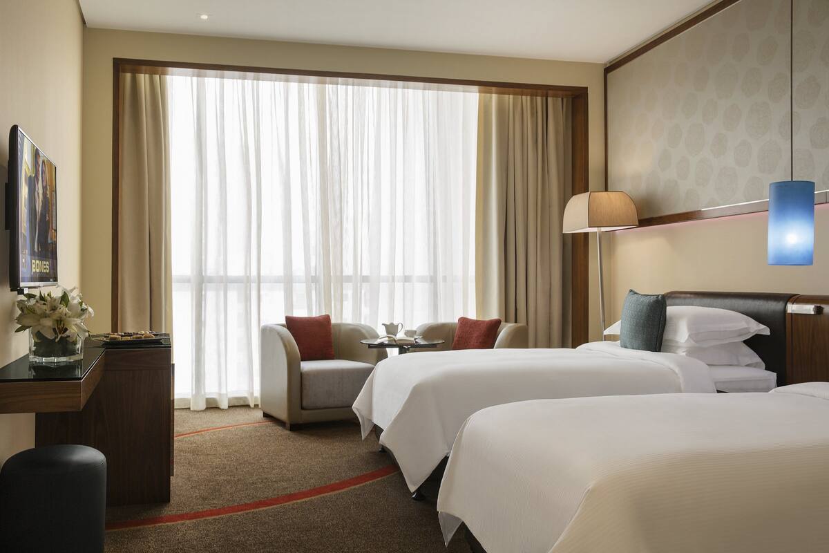 Habitación de hotel con dos camas, un escritorio con lámpara, una silla y un ventanal con cortinas traslúcidas.