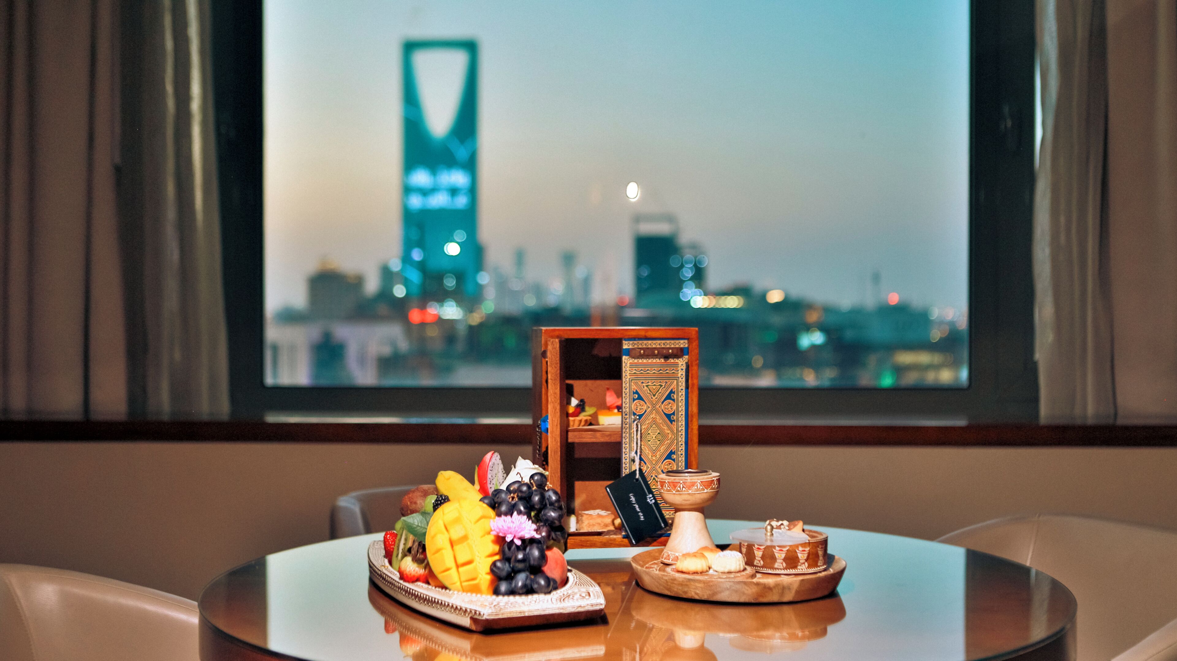 Una mesa con una bandeja de frutas, una caja decorativa y un cuenco pequeño con una vela, frente a una ventana que ofrece vistas de la ciudad.