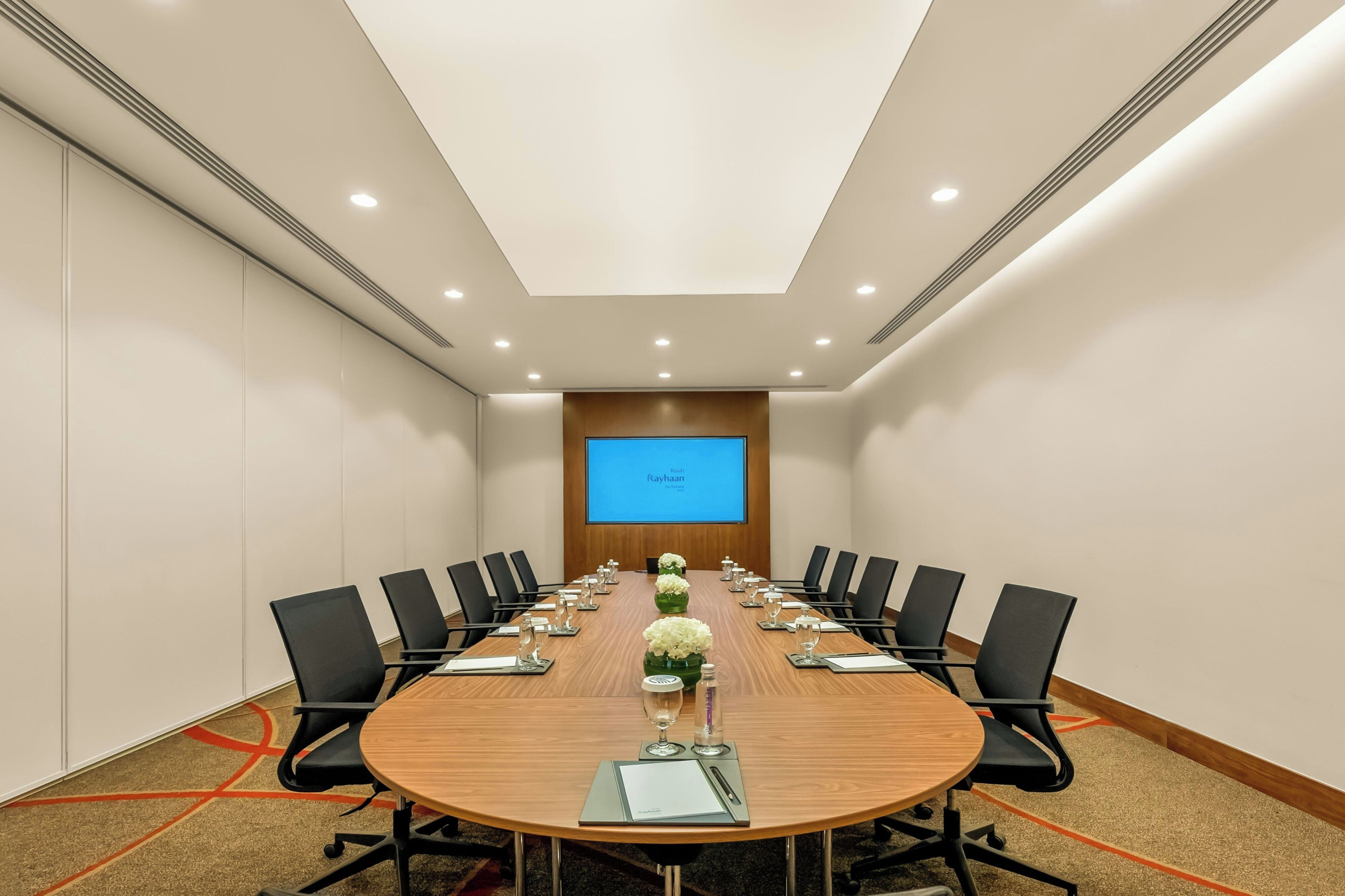 Un salón de conferencias con una larga mesa, sillas negras y una pantalla al frente.