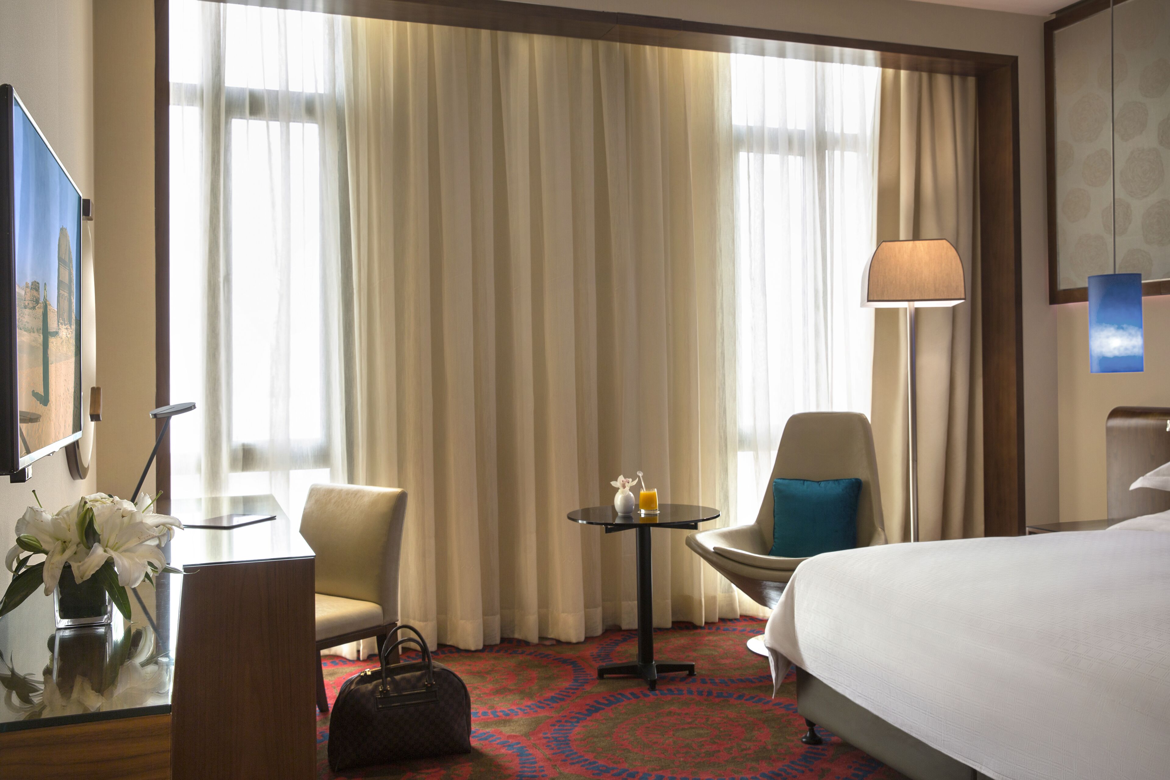 A hotel room with a bed, two chairs, a small table with a lamp, a vase with flowers, and a large window with curtains.