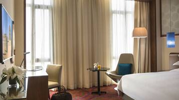 A hotel room with a bed, two chairs, a small table with a lamp, a vase with flowers, and a large window with curtains.