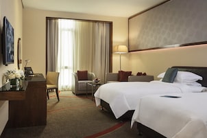 Habitación de hotel con dos camas, un escritorio, una silla, una lámpara y un ventanal grande con cortinas.
