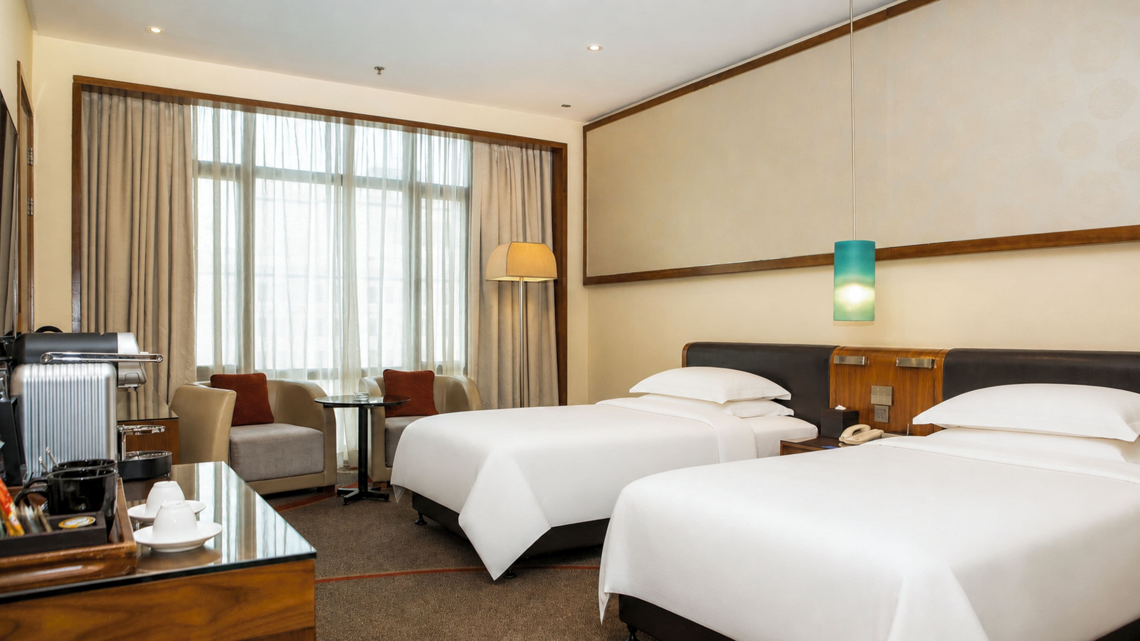 A hotel room with two beds, a desk with a coffee maker, a chair, and a large window with curtains.