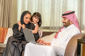 A family of three sitting on a couch with a laptop, in a room with a large window and curtains.