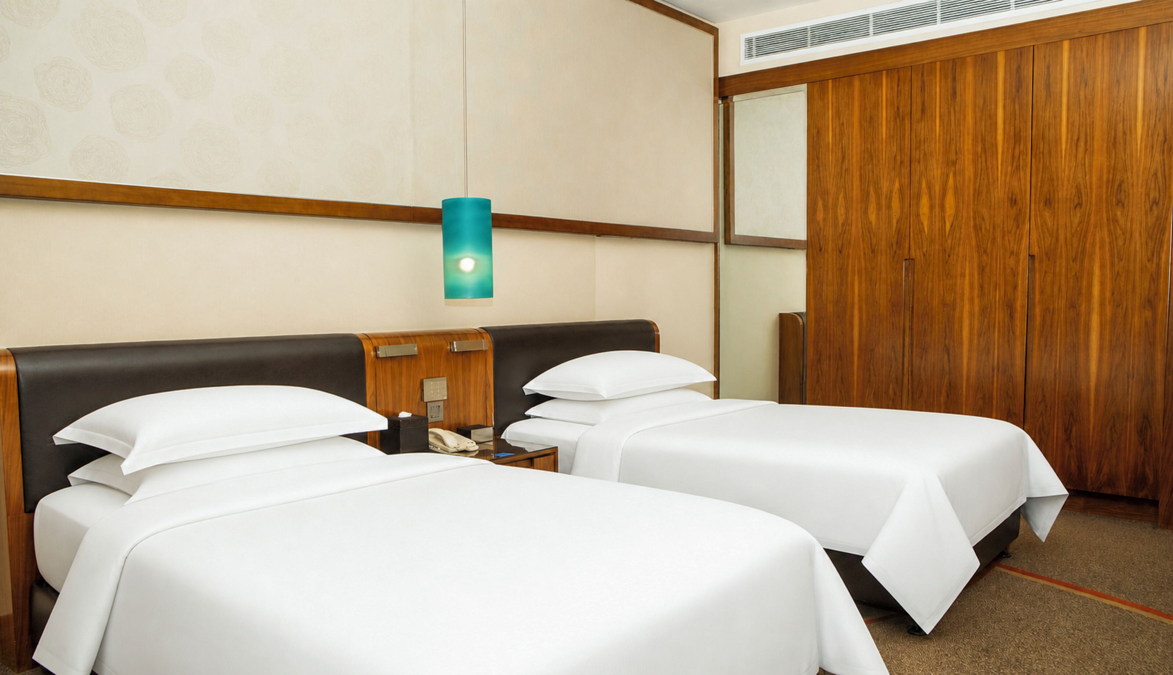 A hotel room with two beds, a wooden wardrobe, a nightstand with a phone, and a wall-mounted light fixture.
