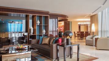 A modern hotel lobby with a curved reception desk, a seating area with a sofa and armchairs, a dining area with a chandelier, and a large TV screen displaying a movie.