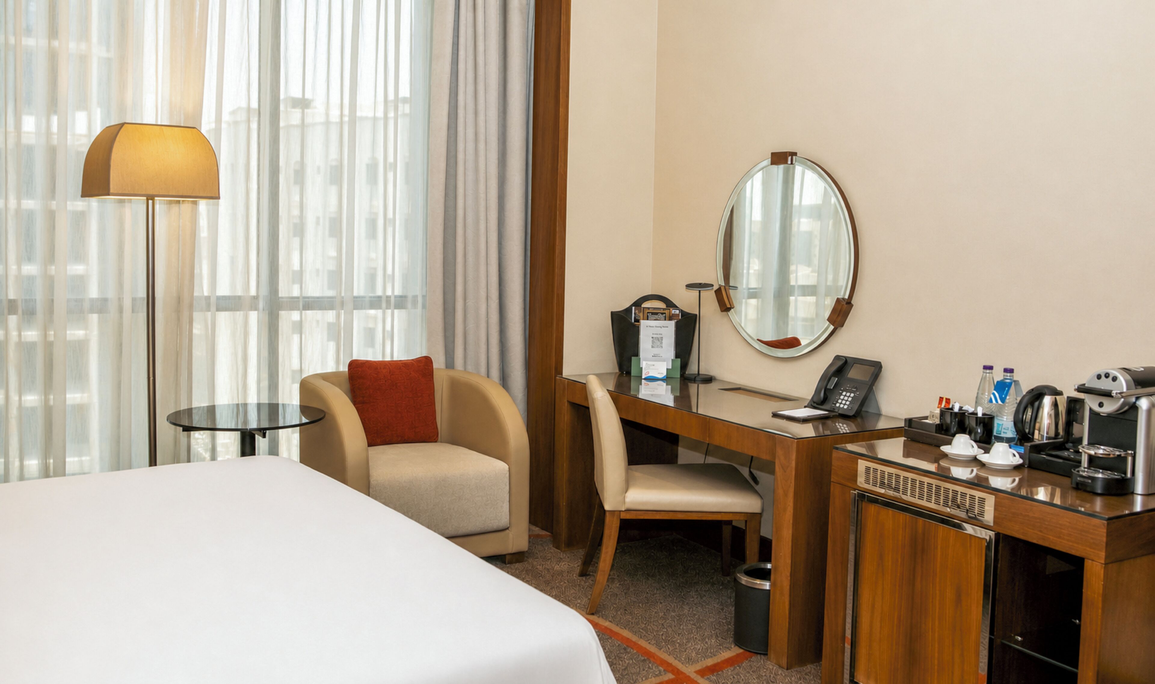 A hotel room with a bed, a desk with a telephone, a chair, a coffee machine, and a large window with curtains.